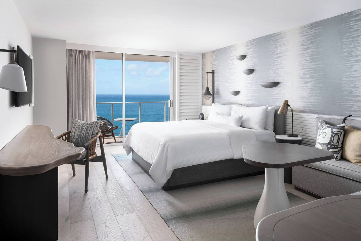 Una habitación de hotel moderna con una cama grande, una mesa de comedor, una silla y vista al océano.