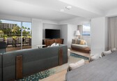 A modern hotel room with a balcony, a flat-screen TV, a sofa, a dining area, and a view of palm trees and buildings.