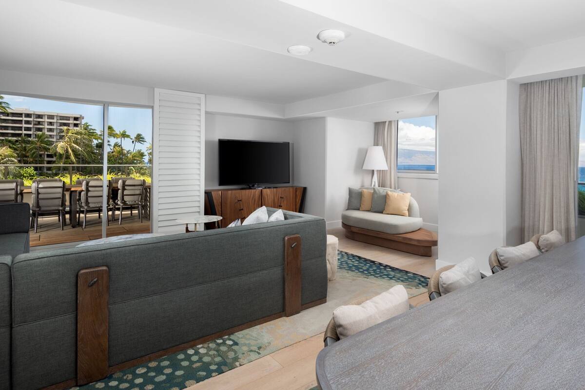 Una habitación de hotel moderna con balcón, televisión de pantalla plana, sofá, zona de comedor y vistas a palmeras y edificios.
