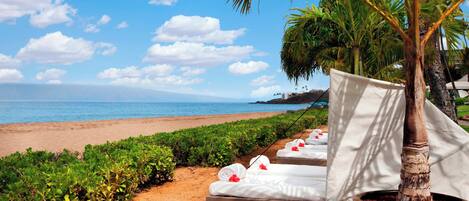 Eine Strandlounge mit weißen Handtüchern und roten Blumen, eine Palme und Blick aufs Meer.