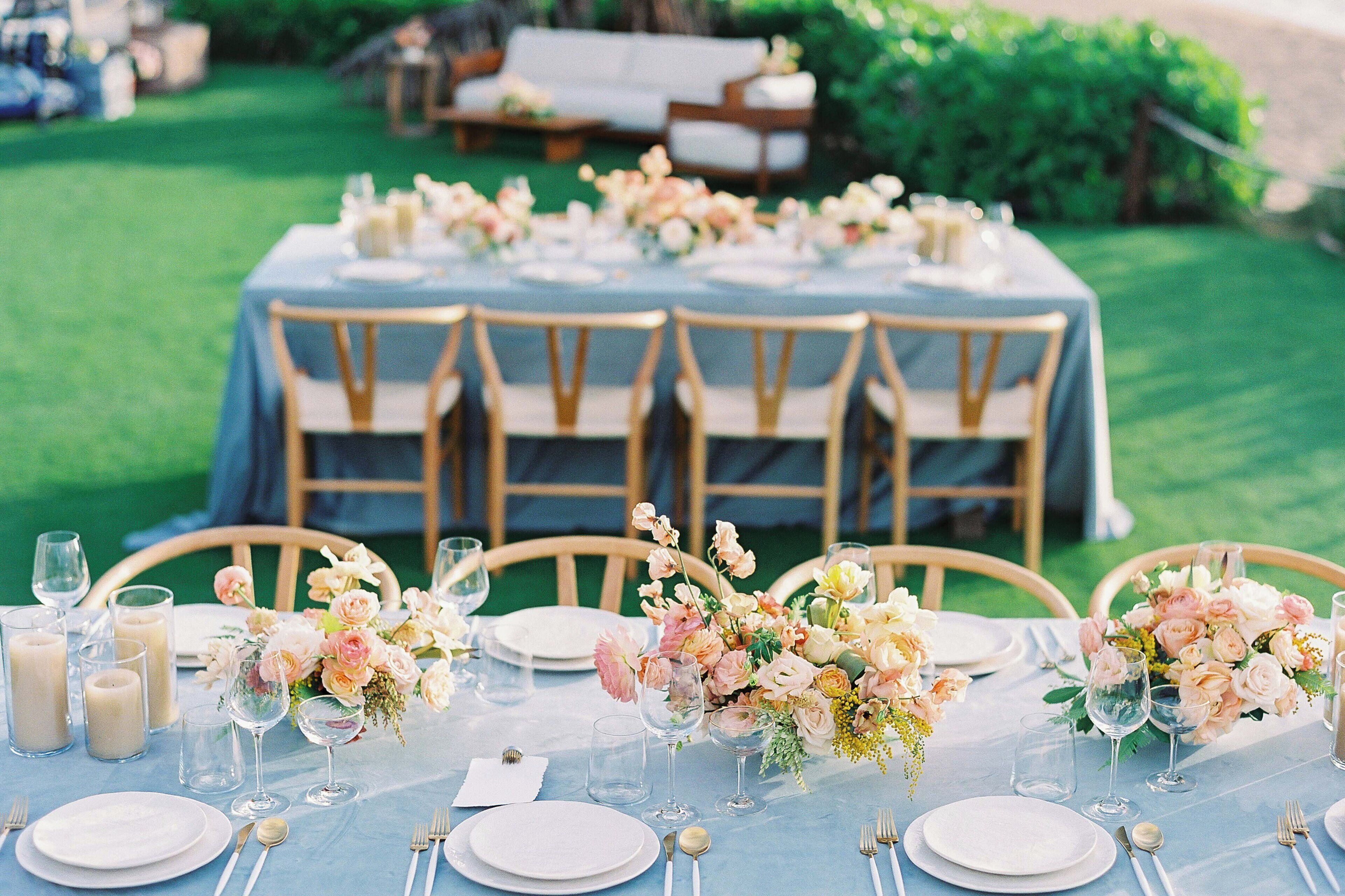 Une table dressée pour un événement formel en plein air, agrémentée de centres de table floraux, d’assiettes blanches et de verres transparents.