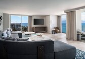 A modern living room with a grey sectional sofa, a flat-screen TV, a small round table, and a balcony with a view of the ocean.