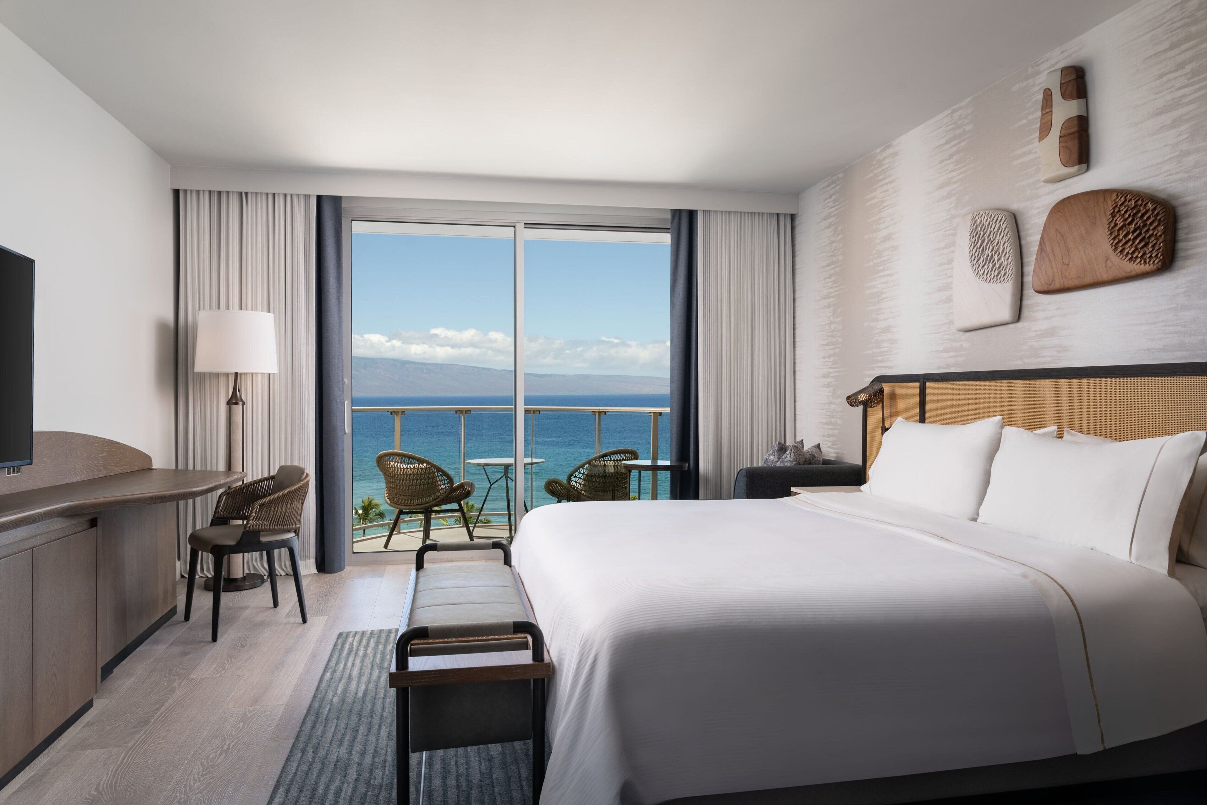 A hotel room with a large bed, a desk, a chair, and a balcony with a view of the ocean.