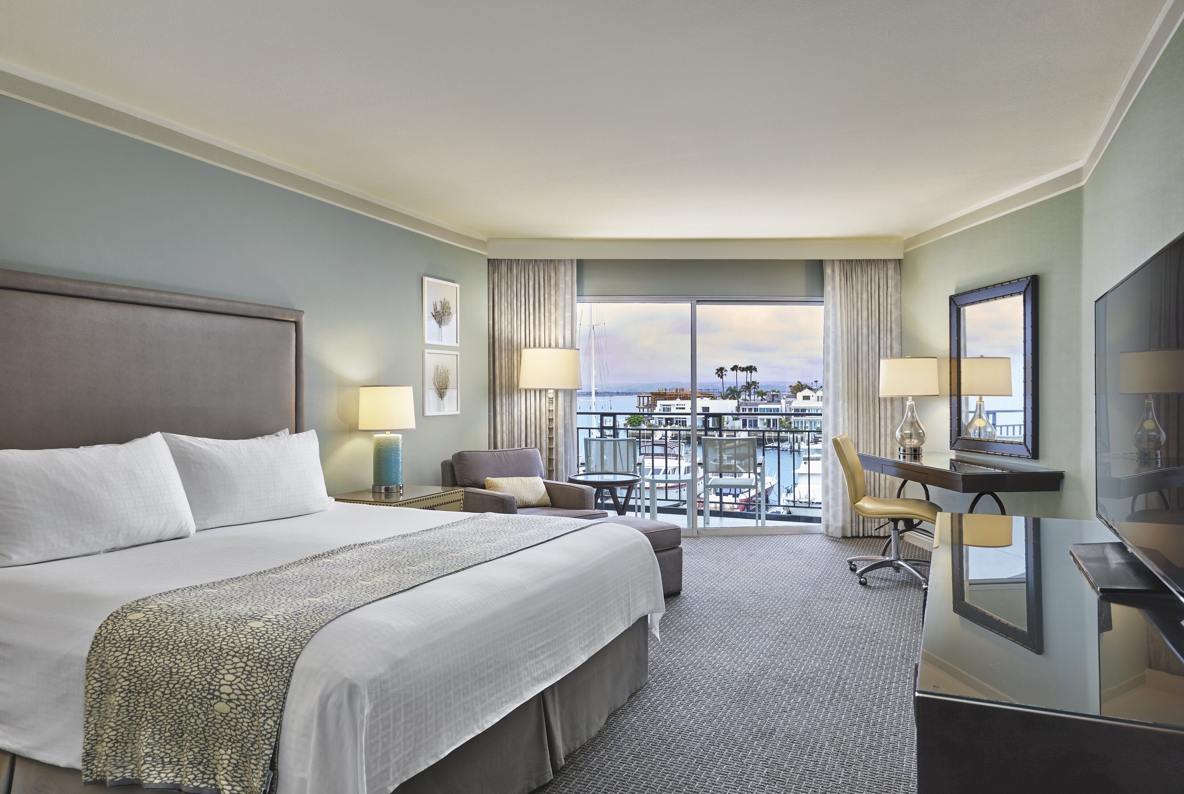 A hotel room with a large bed, a desk, a chair, a TV, and a balcony with a view.