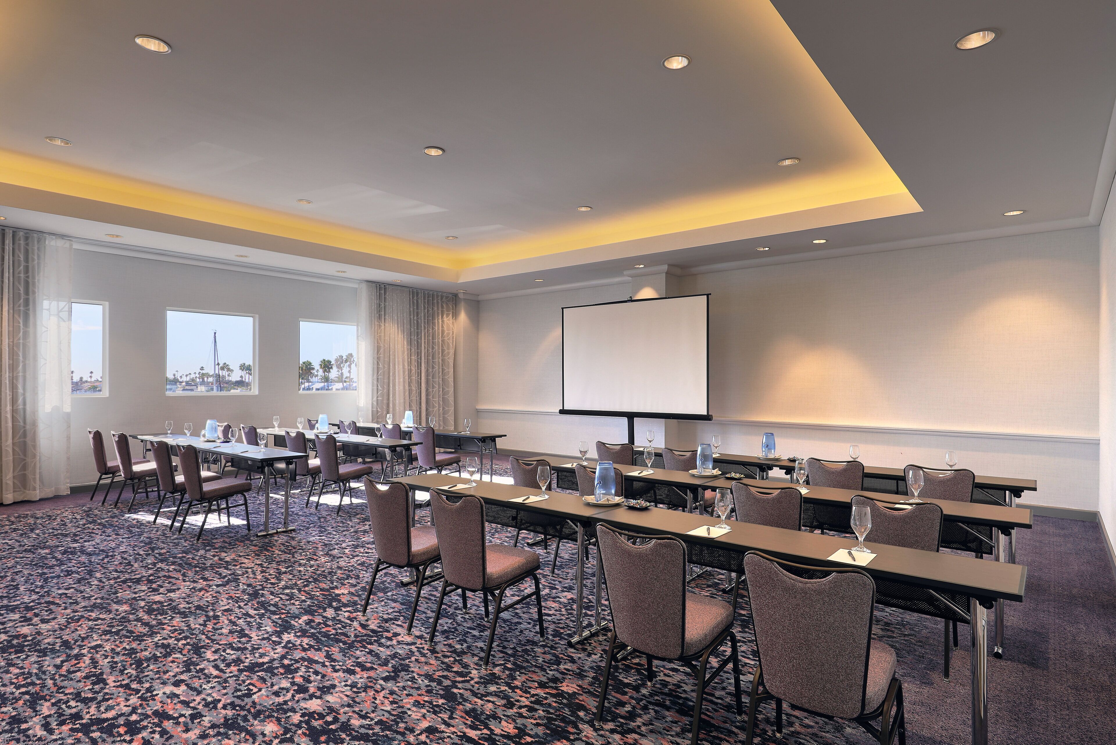 A conference room with tables, chairs, a projector screen, and a city view through large windows.