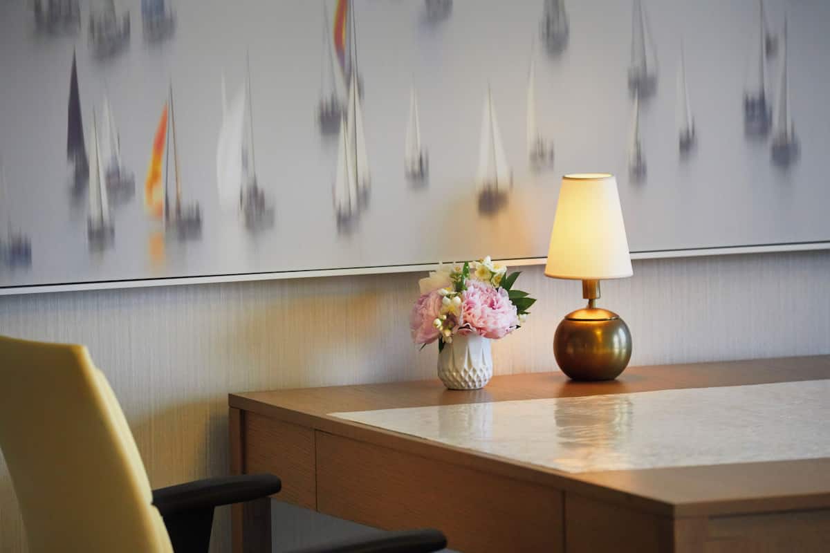 A hotel room with a wooden desk, a vase of flowers, a lamp, and a wall with abstract art.