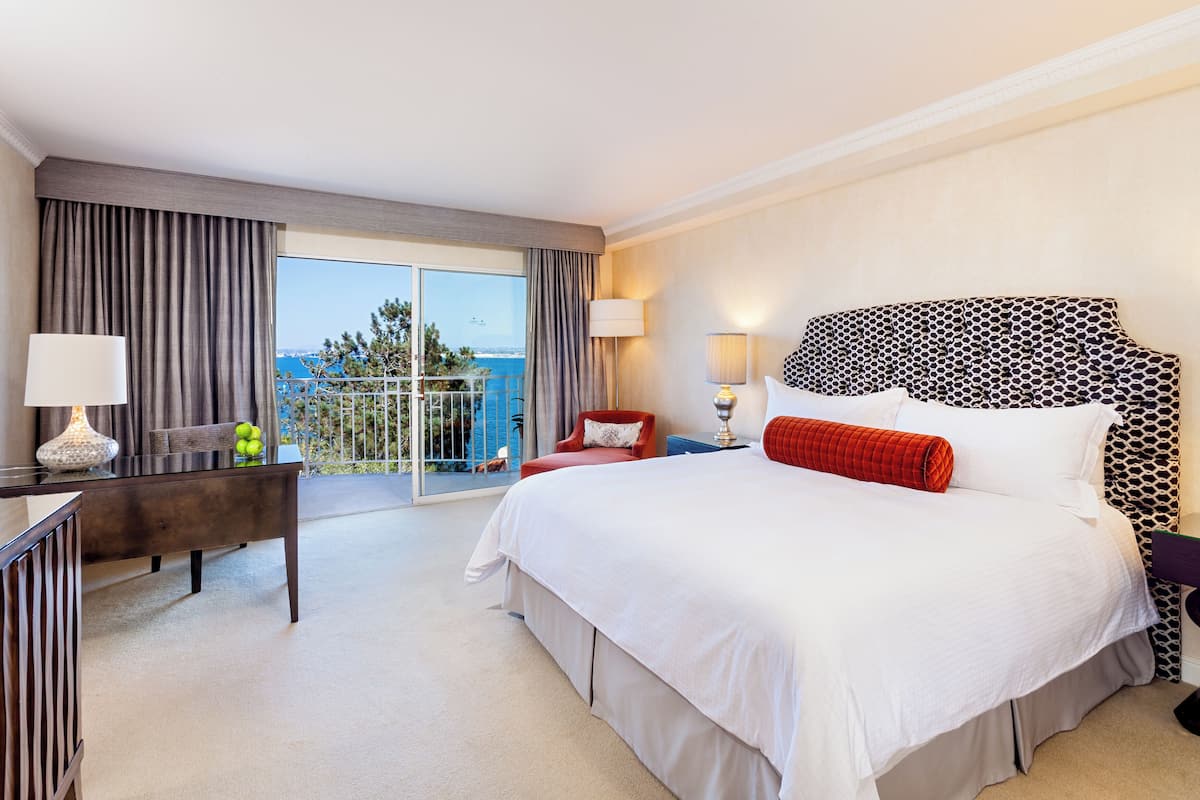 A hotel room with a large bed, a desk, a chair, and a view of the sea through the balcony.