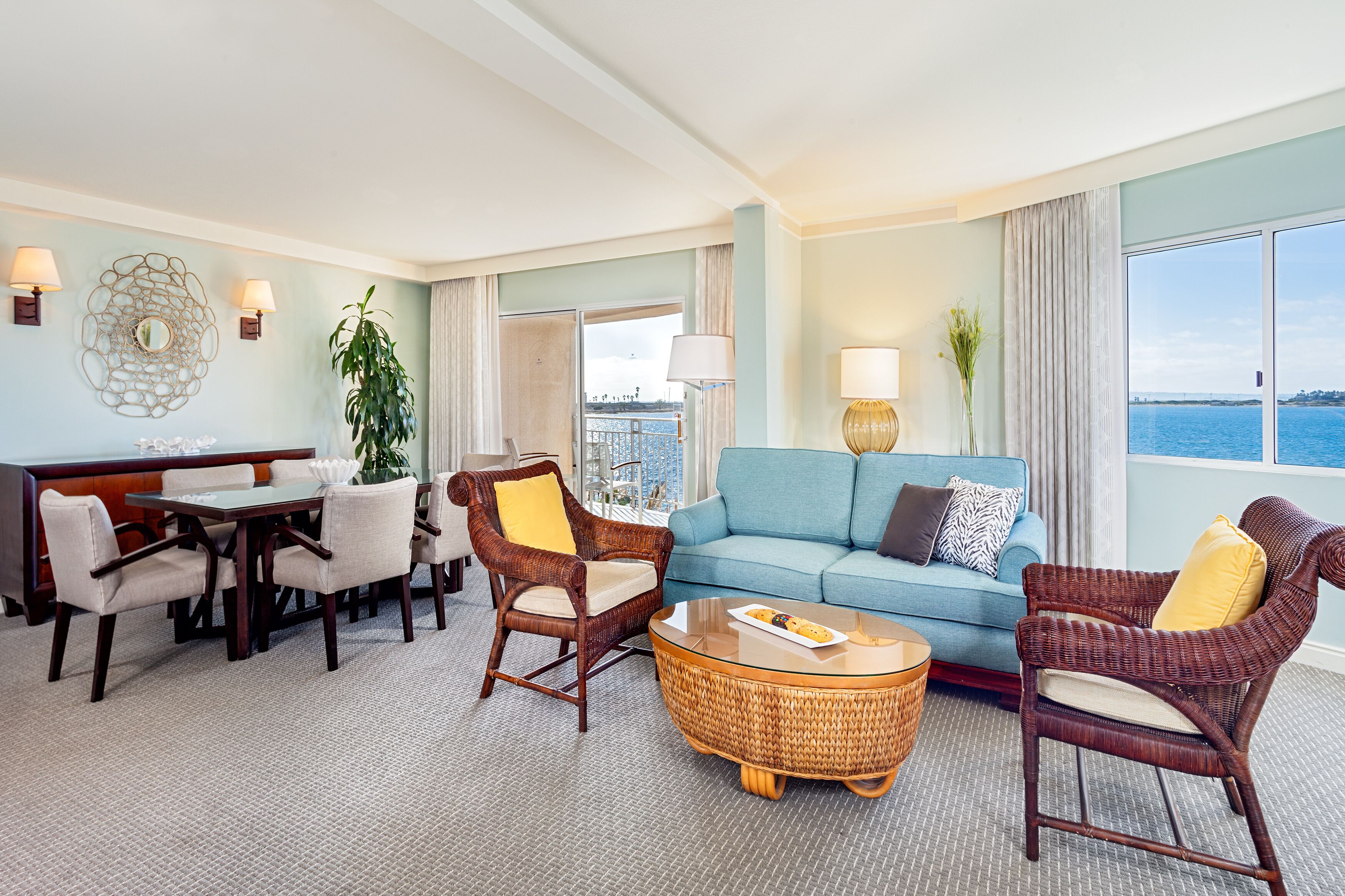 Un salon spacieux comprenant un coin repas, un canapé bleu, des fauteuils en osier et une vue sur la mer.