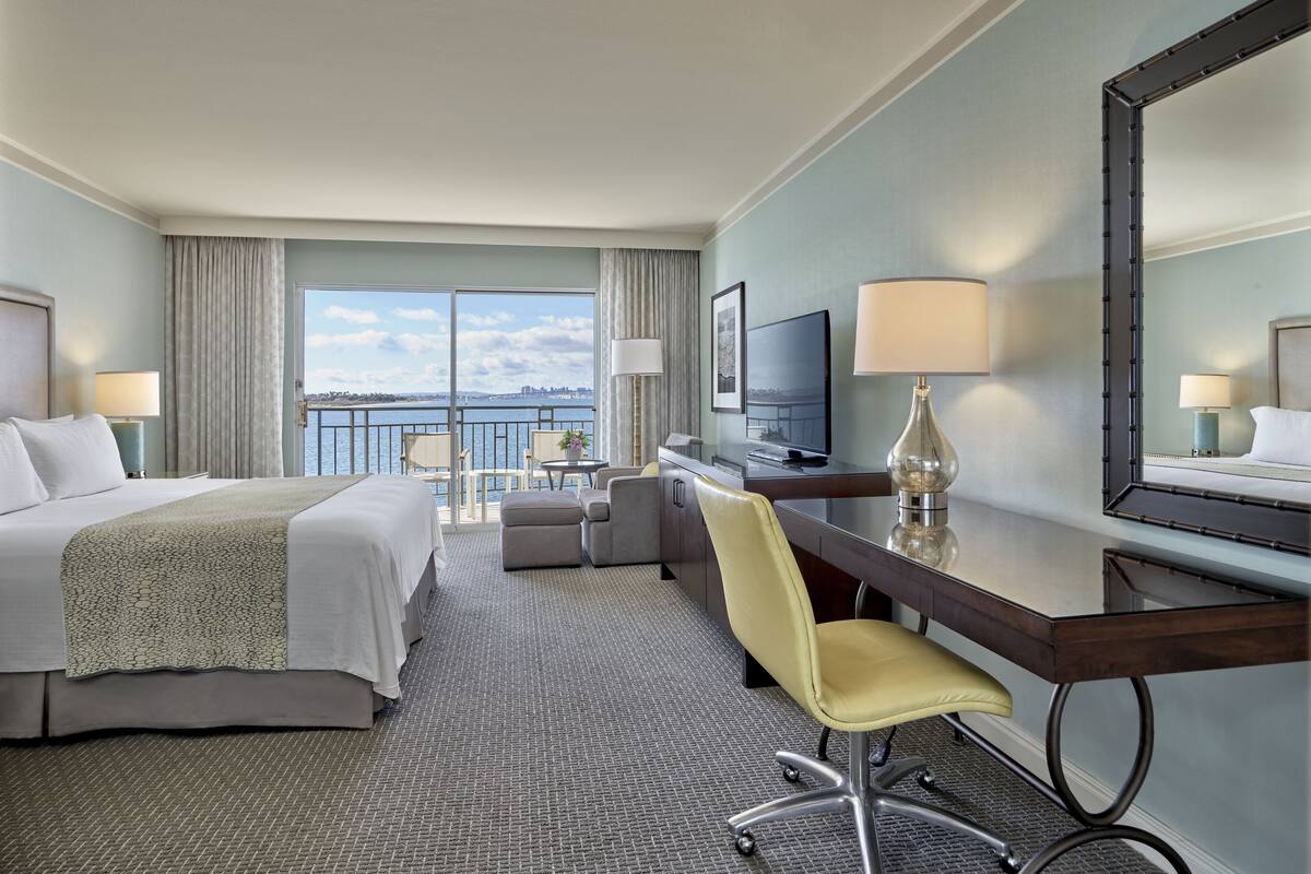 A hotel room with a bed, a desk, a chair, a TV, and a large window with a view of the water.