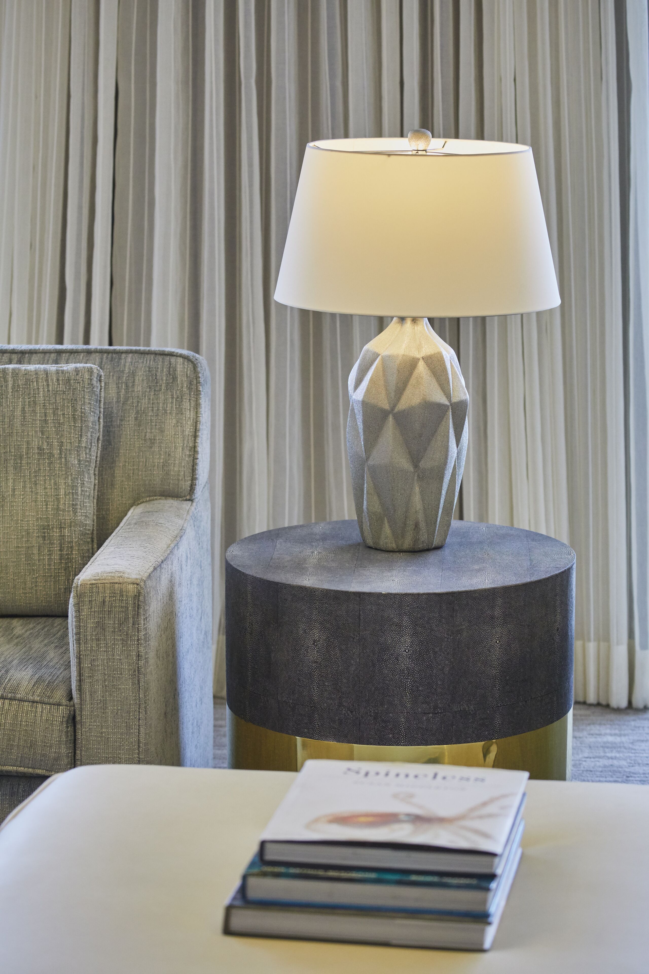 Ein modernes Zimmer mit einem grau gepolsterten Sessel, einem runden Beistelltisch mit einer geometrischen Lampe und einem Stapel Bücher auf einem weißen Pouf.