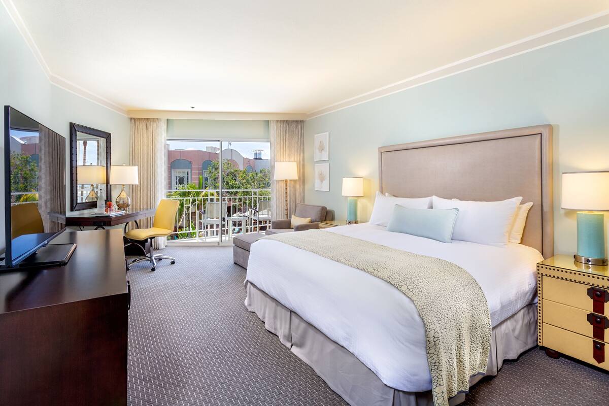 A hotel room with a large bed, a desk with a chair, a TV, and a balcony with outdoor seating.