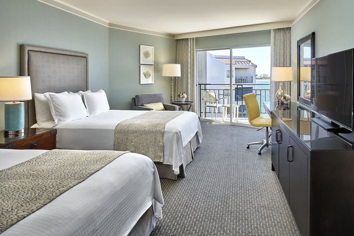 A hotel room with two beds, a TV, a balcony with a view of the sea, and a dining area with chairs and a table.