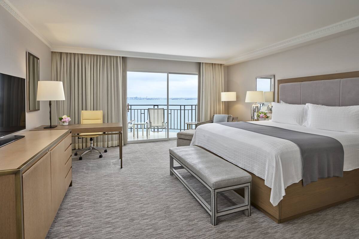 A hotel room with a large bed, a desk with a chair, a bench, and a TV.