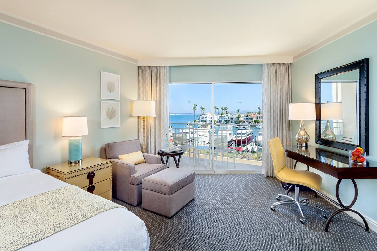 A hotel room with a bed, a sofa, a desk, and a chair. There is a view of the ocean and palm trees outside the window.