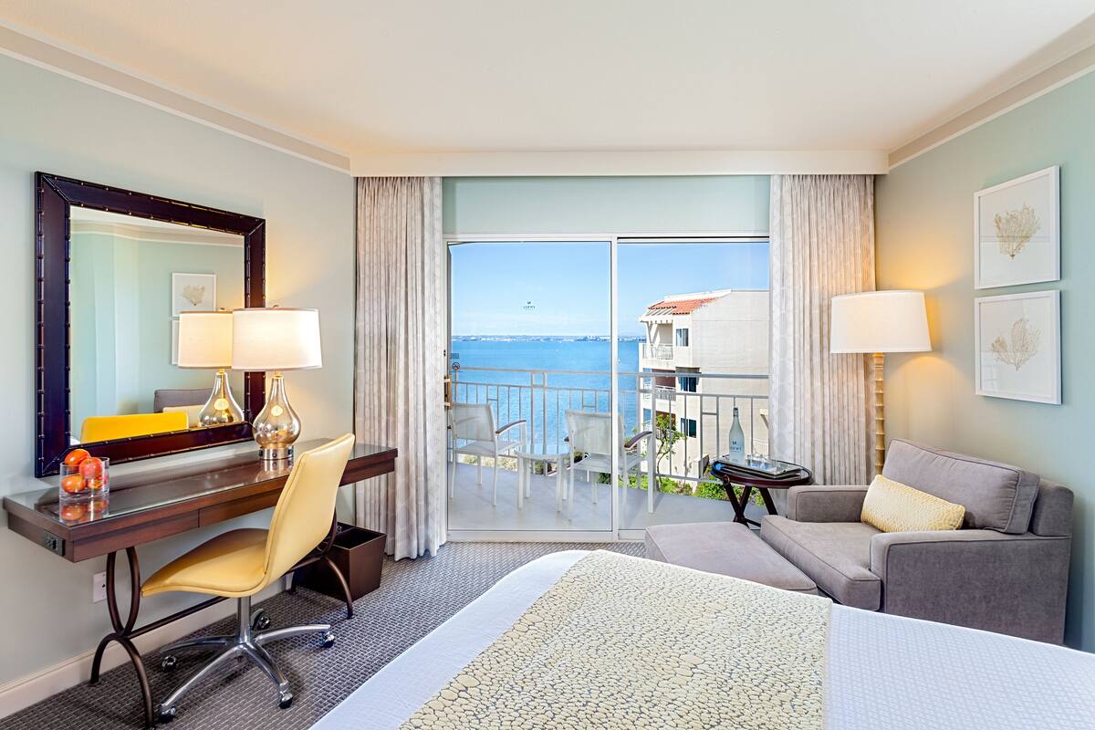 A hotel room with a bed, a desk with a chair, a sofa, a large window with a view of the sea, and a balcony.