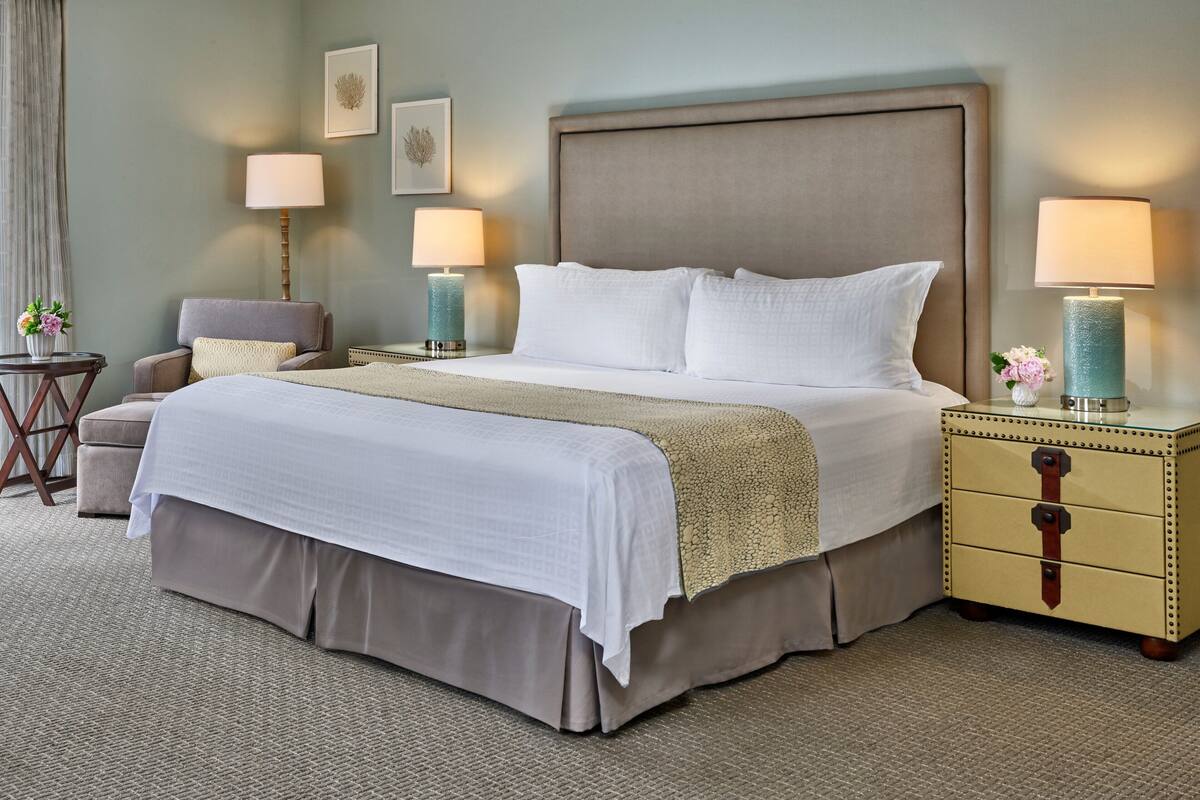 A neatly arranged hotel room with a large bed, two bedside tables with lamps, a chair, and a small table with a vase of flowers.