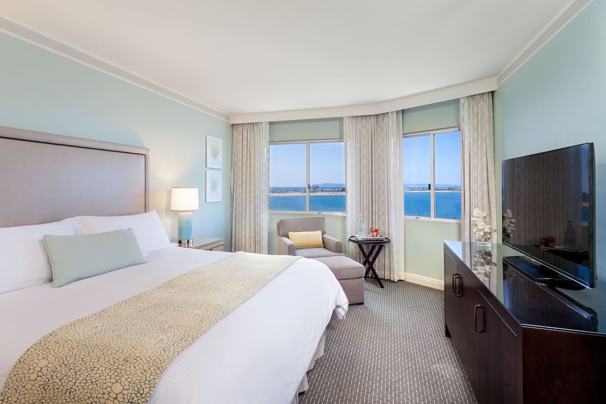 A hotel room with a large bed, a TV, a desk, and a view of the ocean.