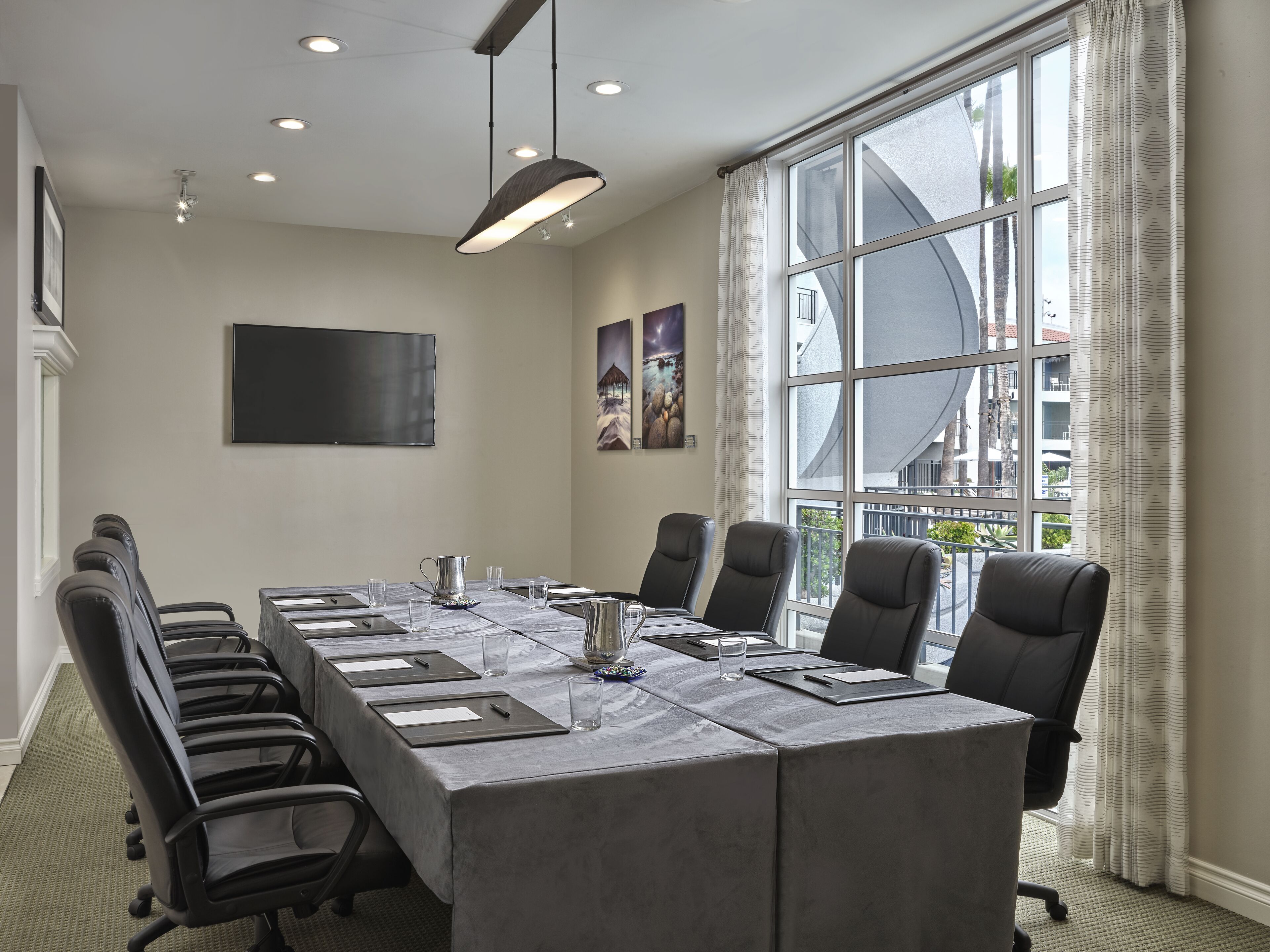 Sala de conferência com uma longa mesa, cadeiras, tela de projetor e grandes janelas com cortinas.