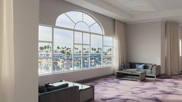 A spacious room with a large arched window overlooking a marina and palm trees. There is a sofa and a coffee table in the foreground.