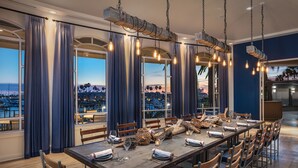 A dining area with a long table set for a meal, large windows with curtains, and hanging light fixtures. - Loews Coronado Bay Resort (Coronado)
