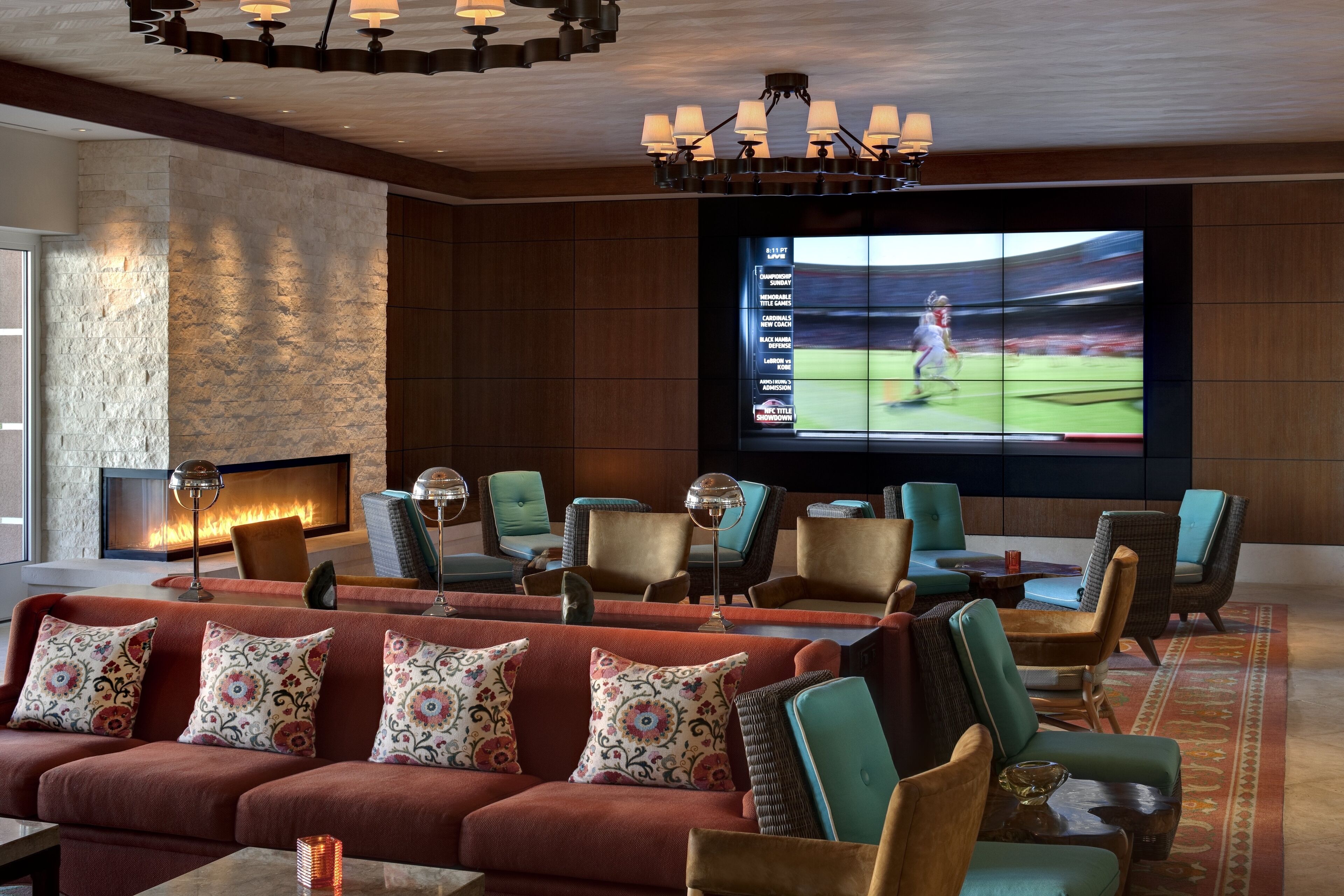 A modern lounge area with a large flat-screen TV displaying a sports game, comfortable seating, a fireplace, and a stone accent wall.