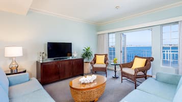 A hotel room with a large window offering a sea view, a television on a wooden cabinet, a wicker coffee table, and two upholstered chairs.