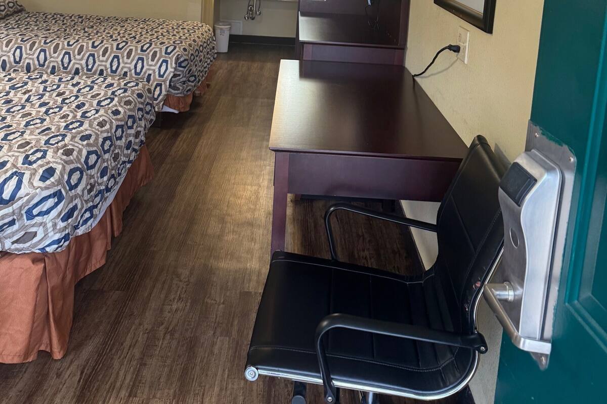 Quarto de hotel com cama, escrivaninha, cadeira, espelho e banheiro ao fundo.