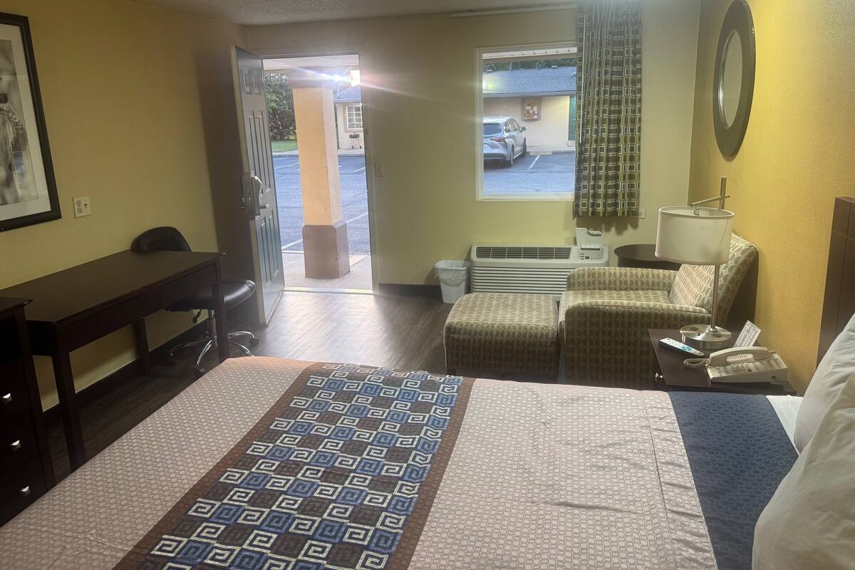 Quarto de hotel com cama, escrivaninha, poltrona, sofá, janela com vista para o estacionamento e ar condicionado.