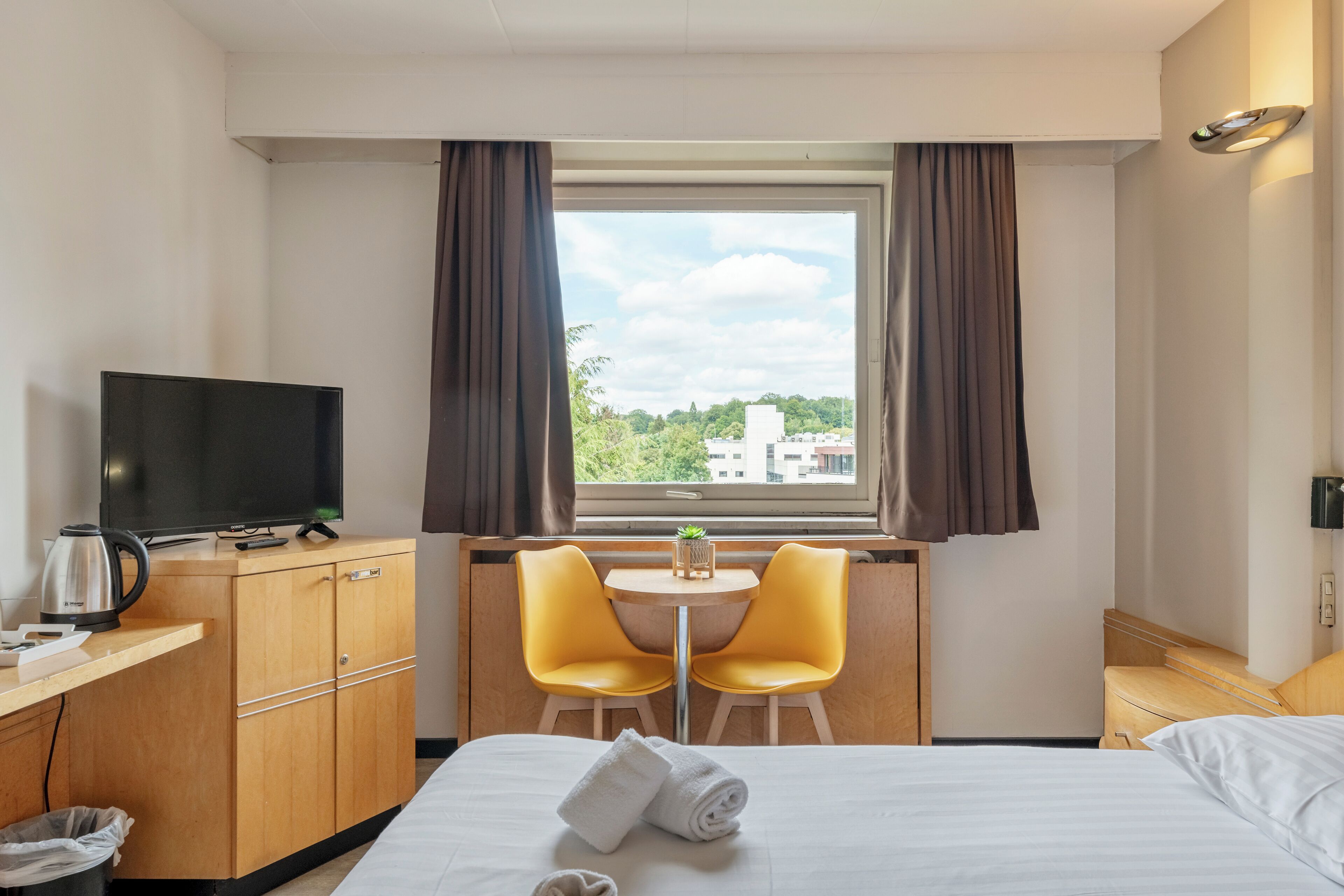 A hotel room with a bed, a TV, a desk with a chair, and a window with curtains.