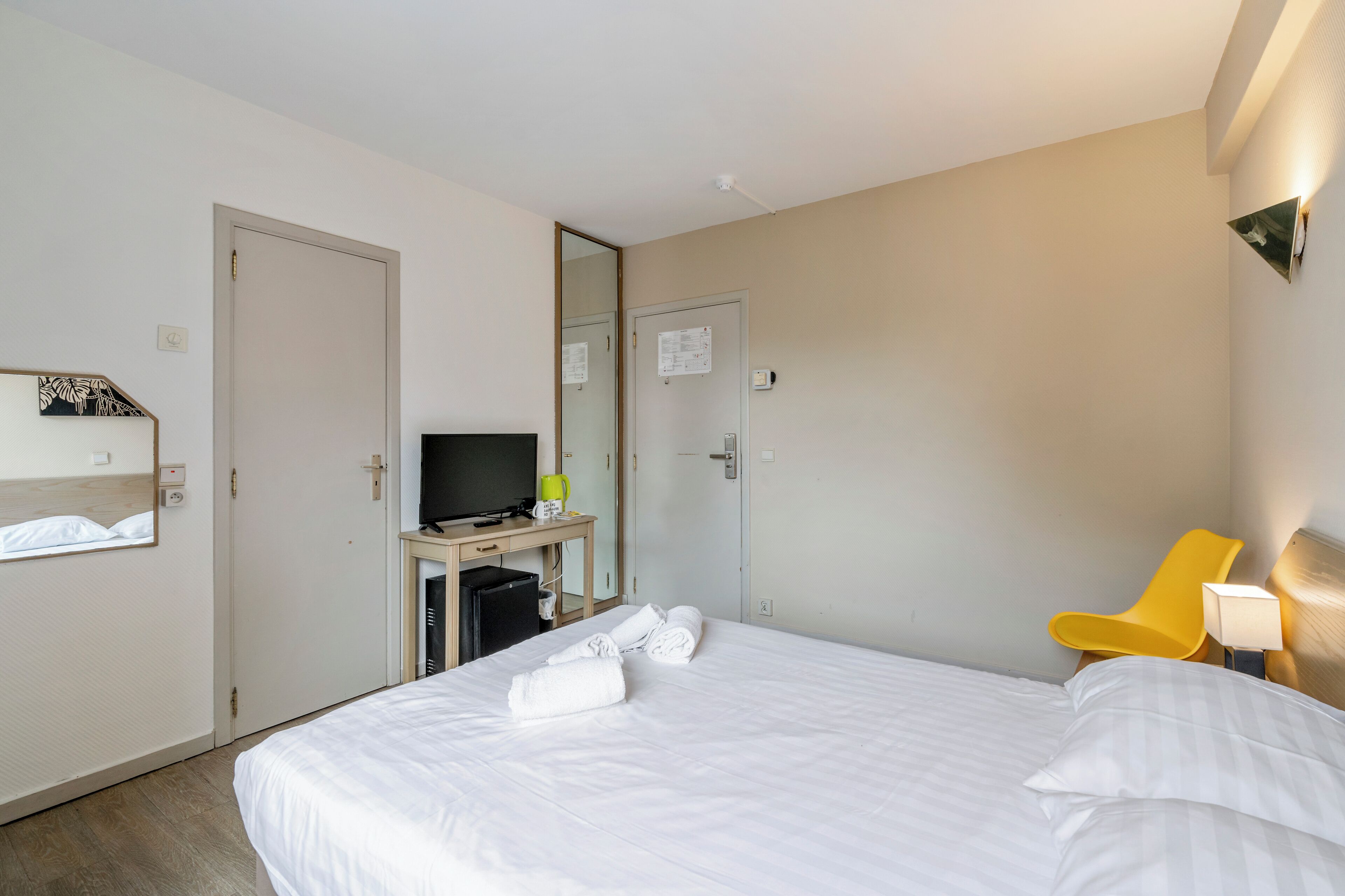 Habitación de hotel con cama, escritorio con computadora, televisor, una silla amarilla y espejo.