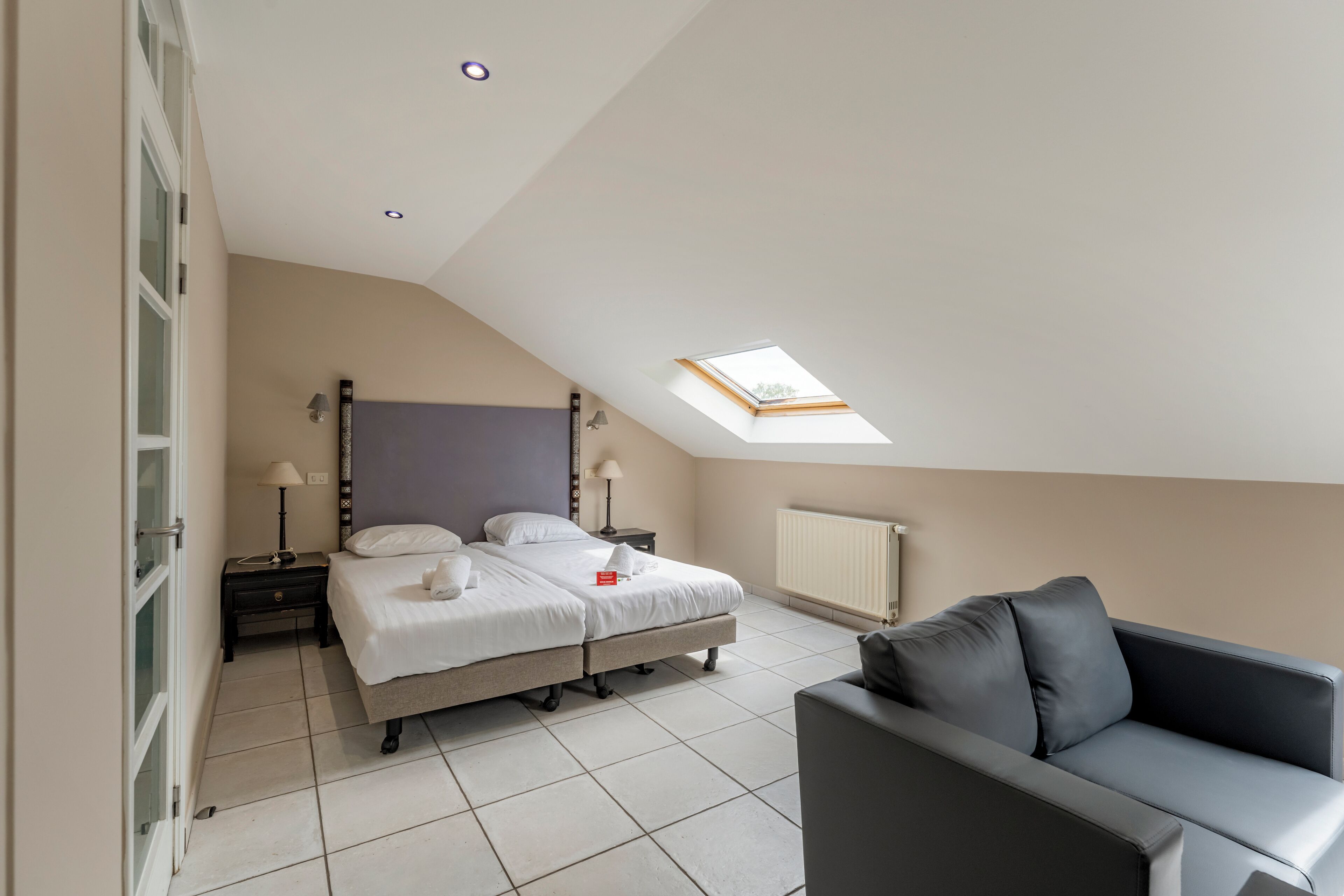 A bedroom with a bed, bedside tables, a nightstand, a sofa, a skylight, and a radiator.