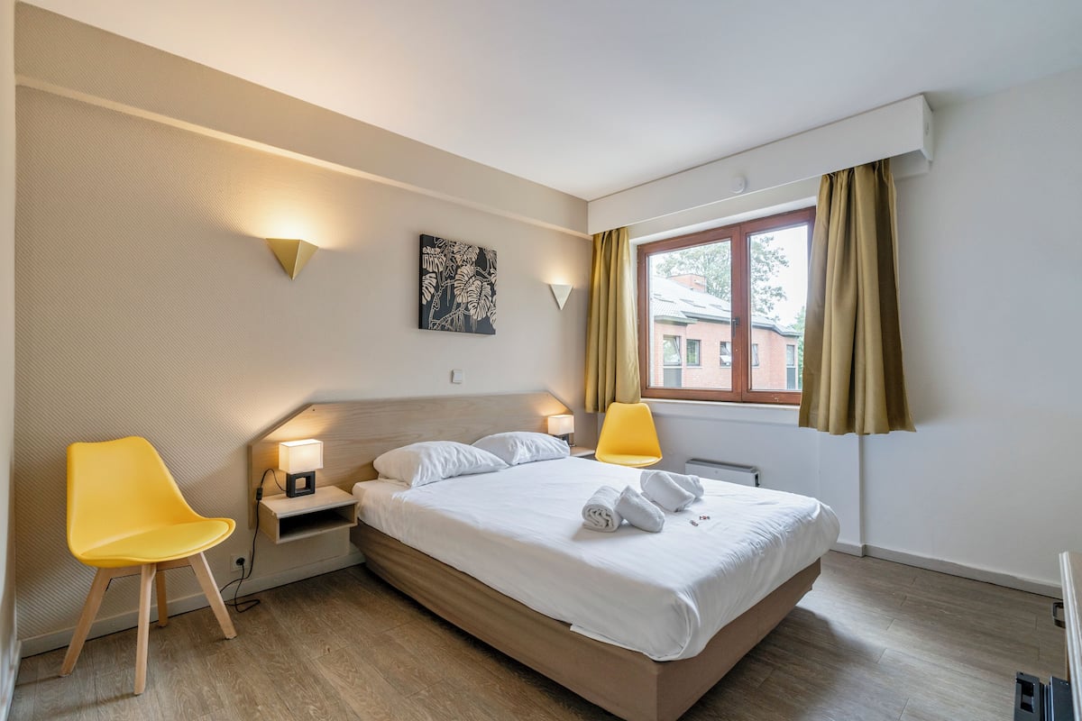 A hotel room with a bed, two bedside lamps, a chair, a small table, a wall-mounted artwork, and a window with curtains.
