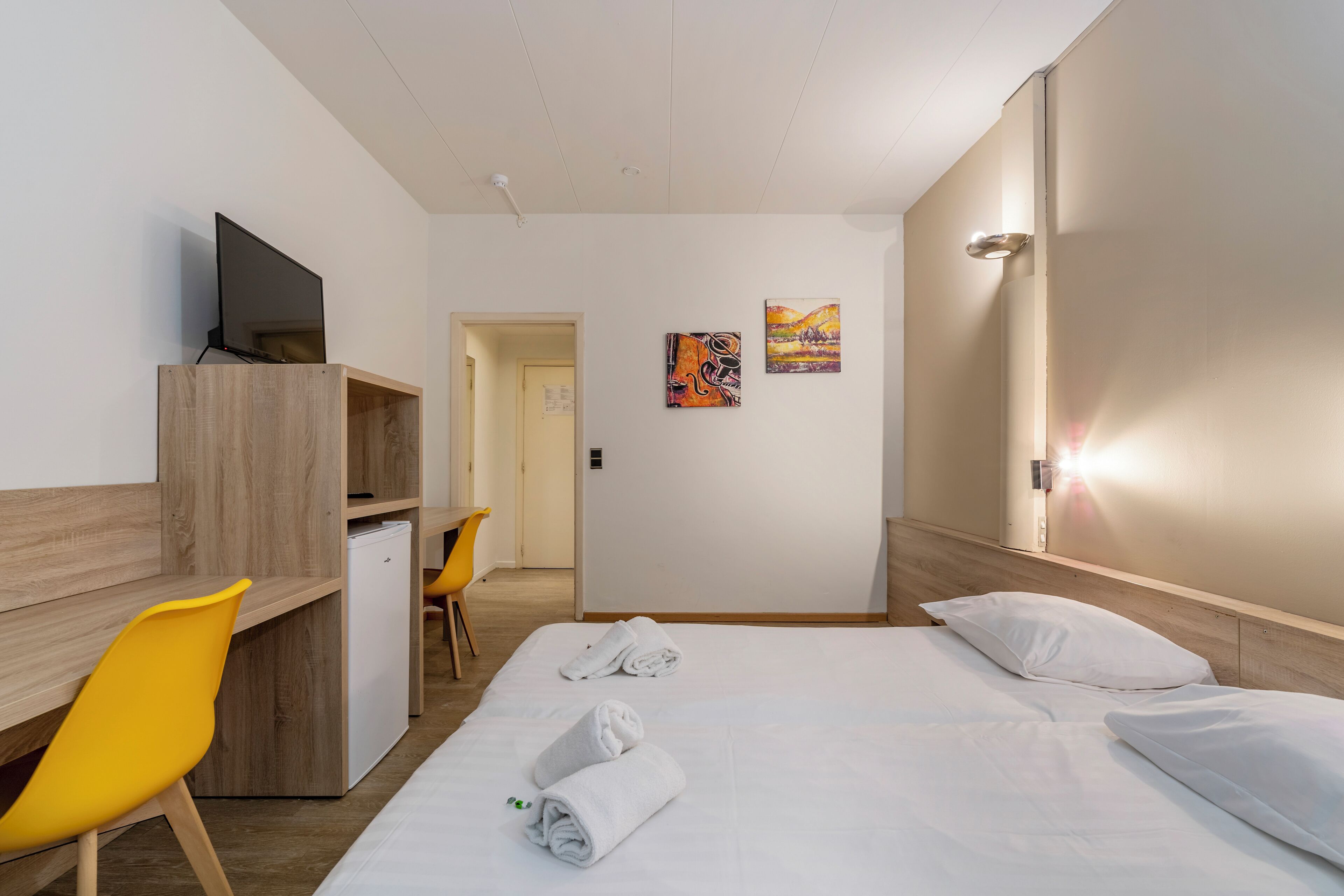 Une chambre d’hôtel équipée d’un lit, d’un bureau, d’une chaise, d’une télévision et d’œuvres d’art accrochées au mur.