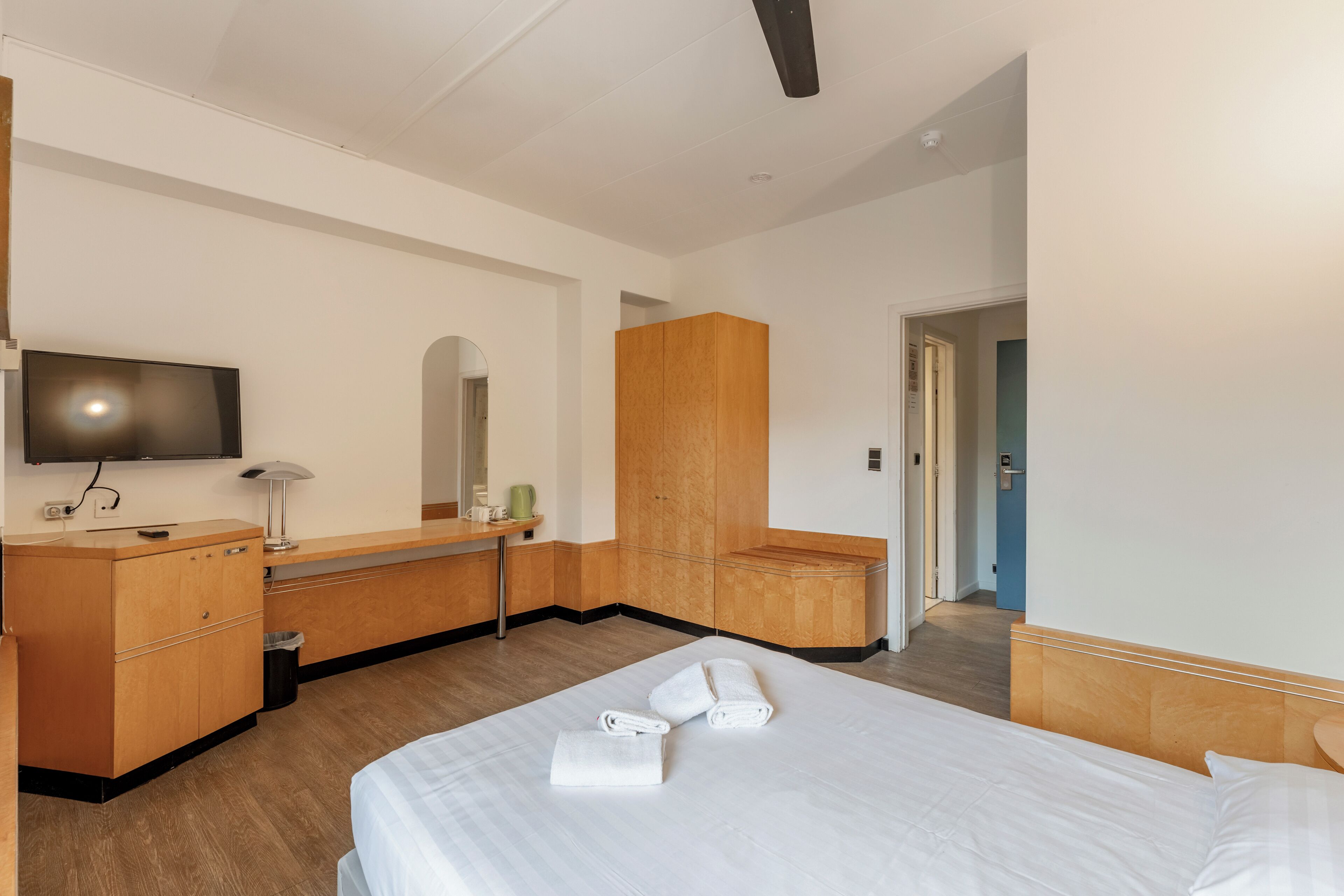 Een hotelkamer met een bed, een houten garderobe, een bureau met een lamp en een televisie aan de muur.