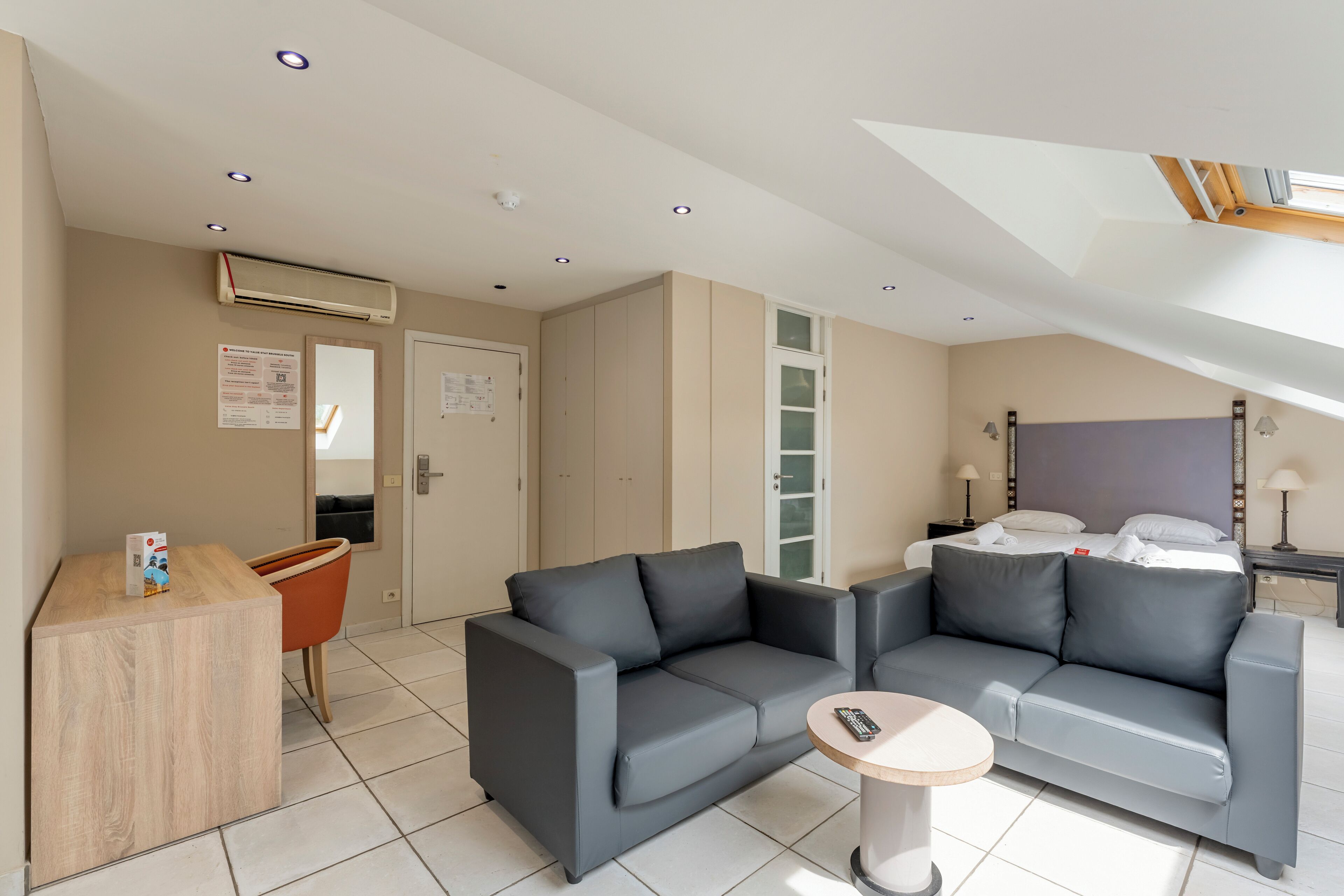 Une chambre d’hôtel moderne avec un canapé gris, une petite table ronde et un lit recouvert de draps blancs.