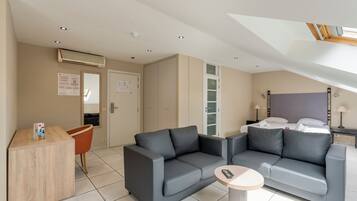 Une chambre d’hôtel moderne avec un canapé gris, une petite table ronde et un lit recouvert de draps blancs.