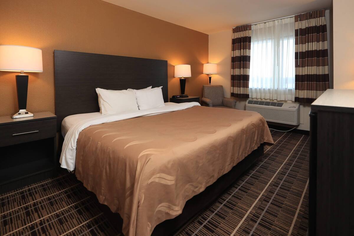 A hotel room with a large bed, two bedside lamps, a chair, an air conditioning unit, and a window with curtains.