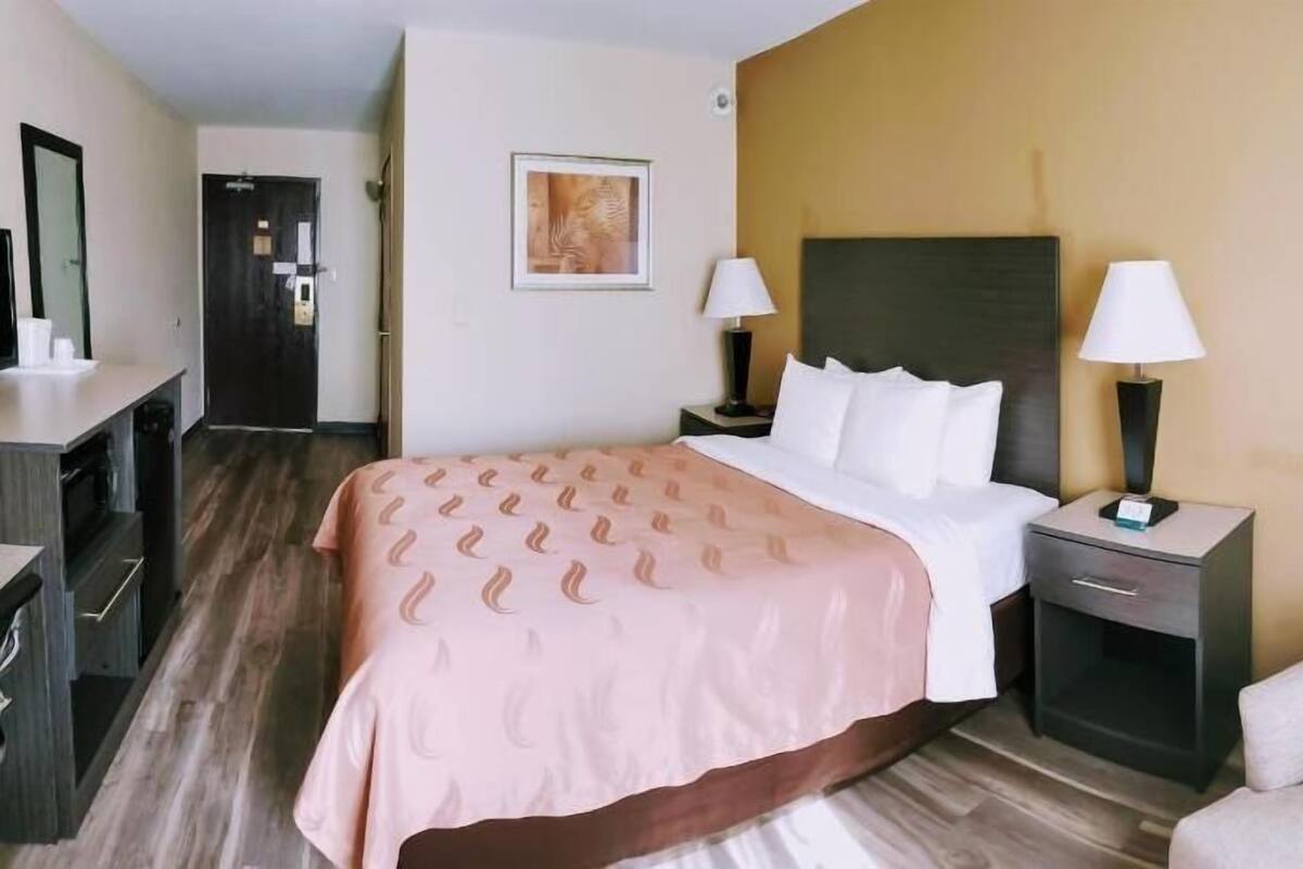 A hotel room with a bed, bedside tables, a desk, a chair, and a painting on the wall.