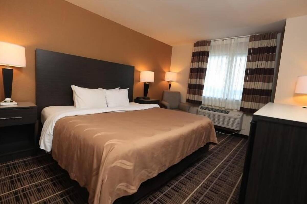 A hotel room with a bed, two bedside tables with lamps, a desk with a chair, and a window with curtains.