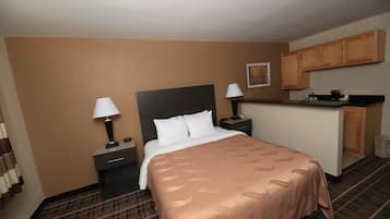 A hotel room with a bed, a nightstand, a lamp, a small desk, a kitchenette, and a trash can.