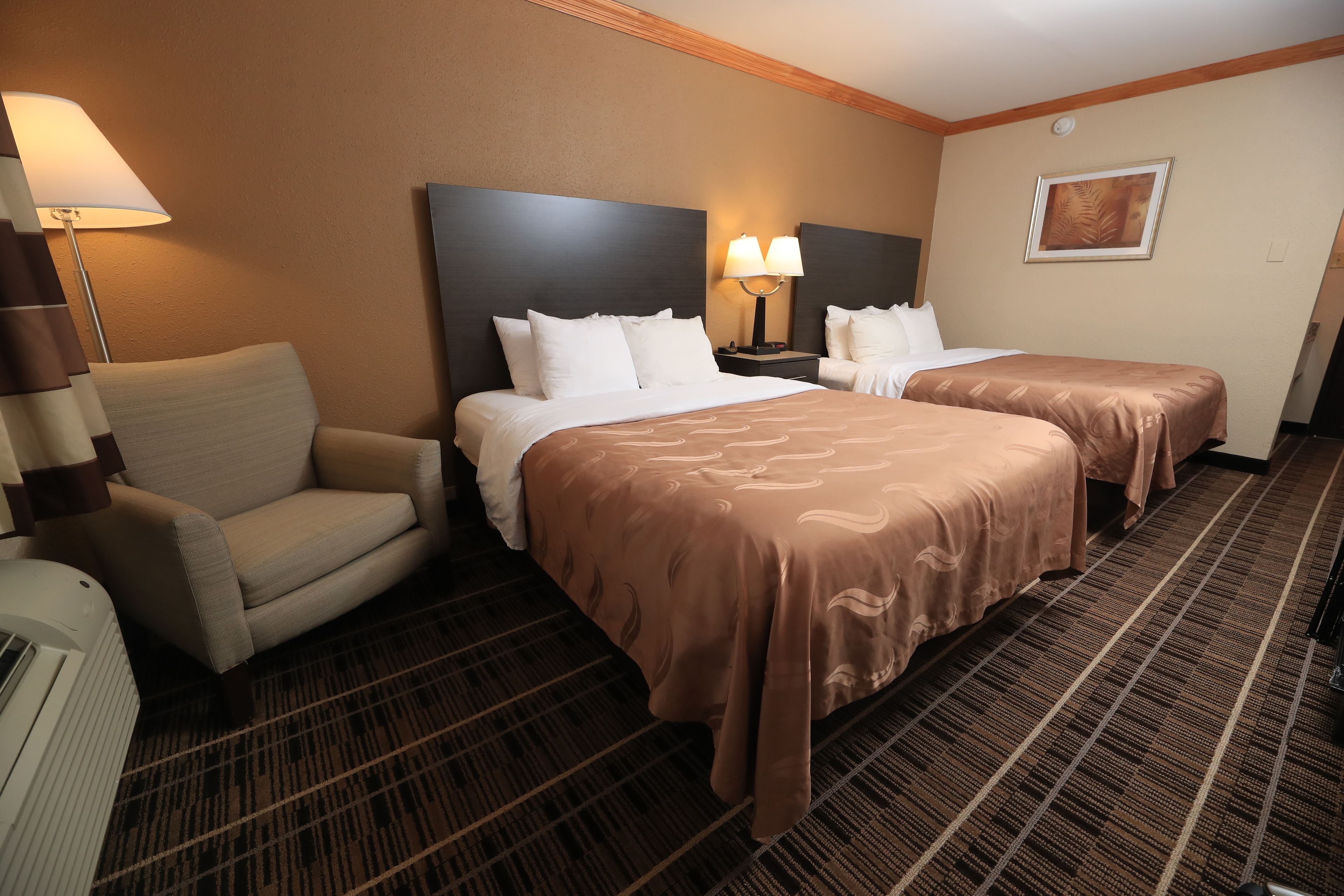 A hotel room with two beds, a chair, a lamp, and a picture on the wall.