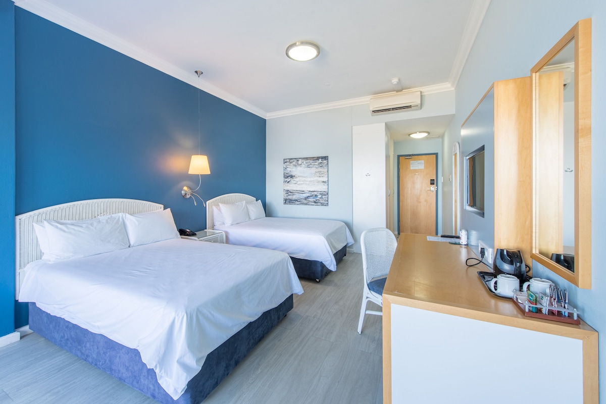 A hotel room with two beds, a desk with a mirror, a chair, and a wall-mounted lamp.