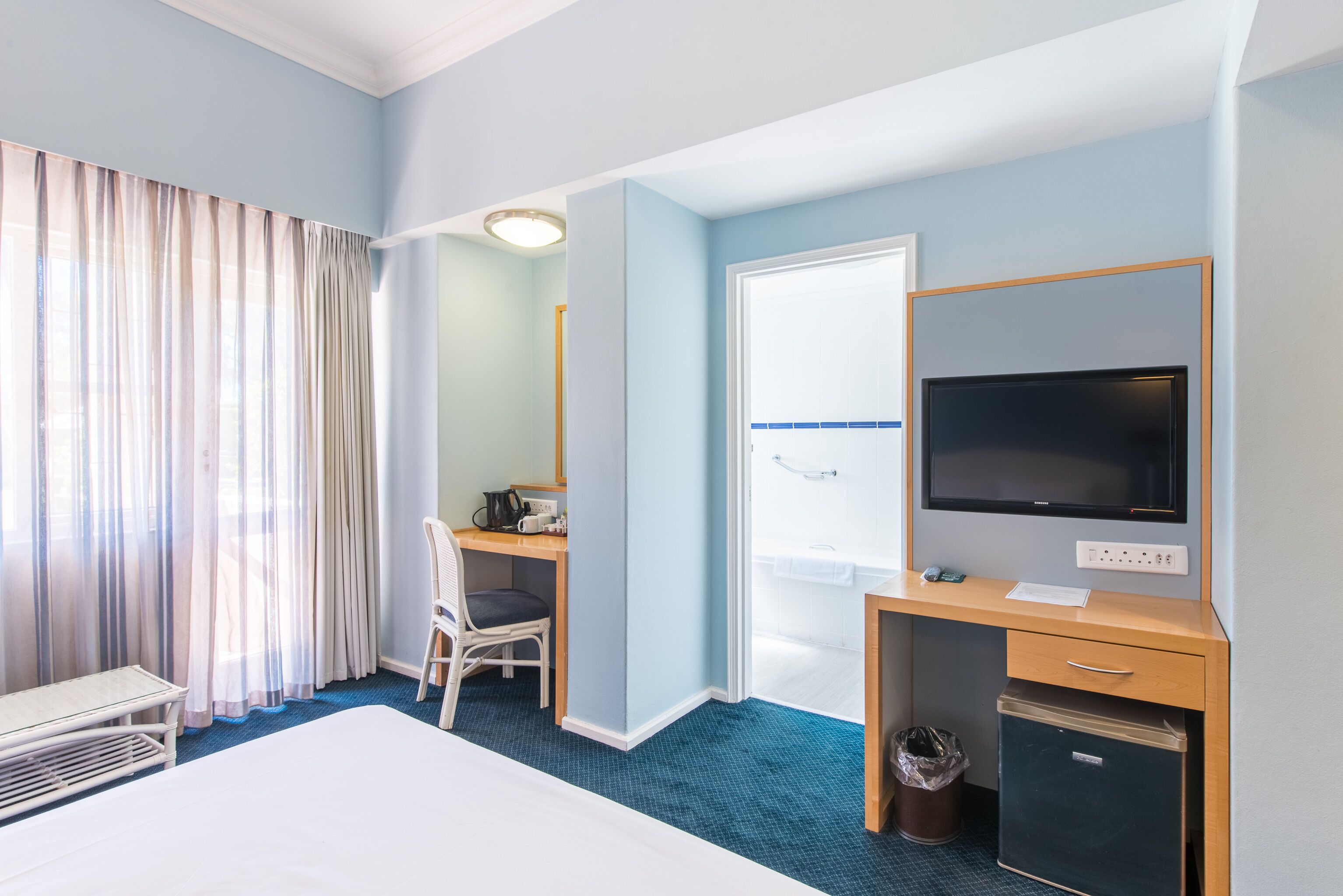 Una habitación de hotel con cama, escritorio con computadora, televisor, silla, basurero y ventana con cortinas.