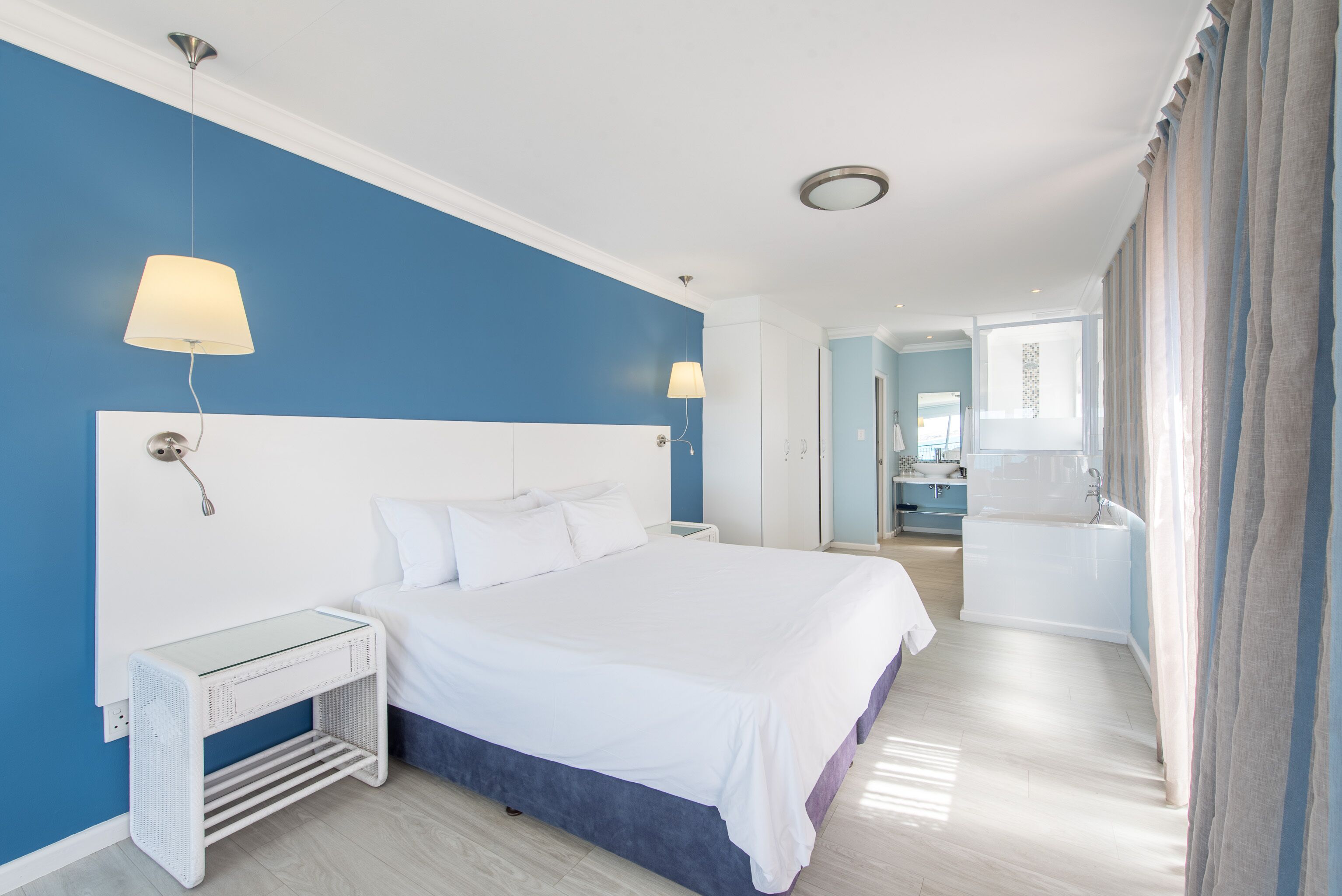 Moderni hotellihuone, jossa on suuri sänky, yöpöytälamput, pieni pöytä ja kylpyhuone, joka näkyy avoimesta ovesta.