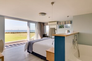 A modern hotel room with a large bed, a bathroom with a bathtub, a desk, and a view of the sea.
