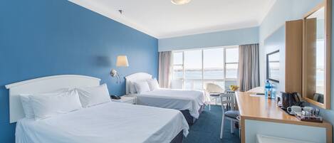 A hotel room with two beds, a desk, a chair, a large window, and a view of the sea.