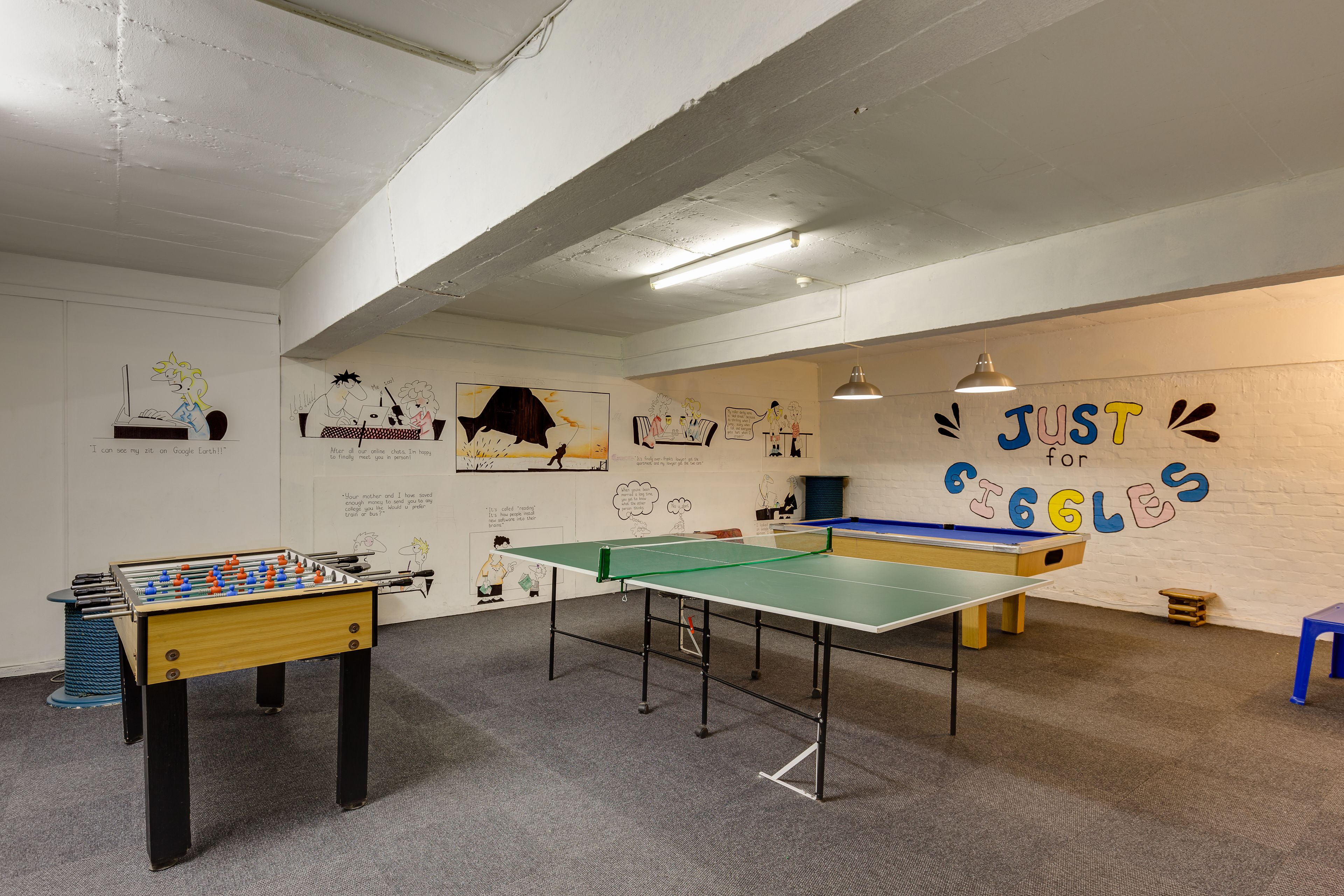 A room with a foosball table, ping pong table, and a pool table. There are drawings on the walls and a sign that says "JUST for giggles".