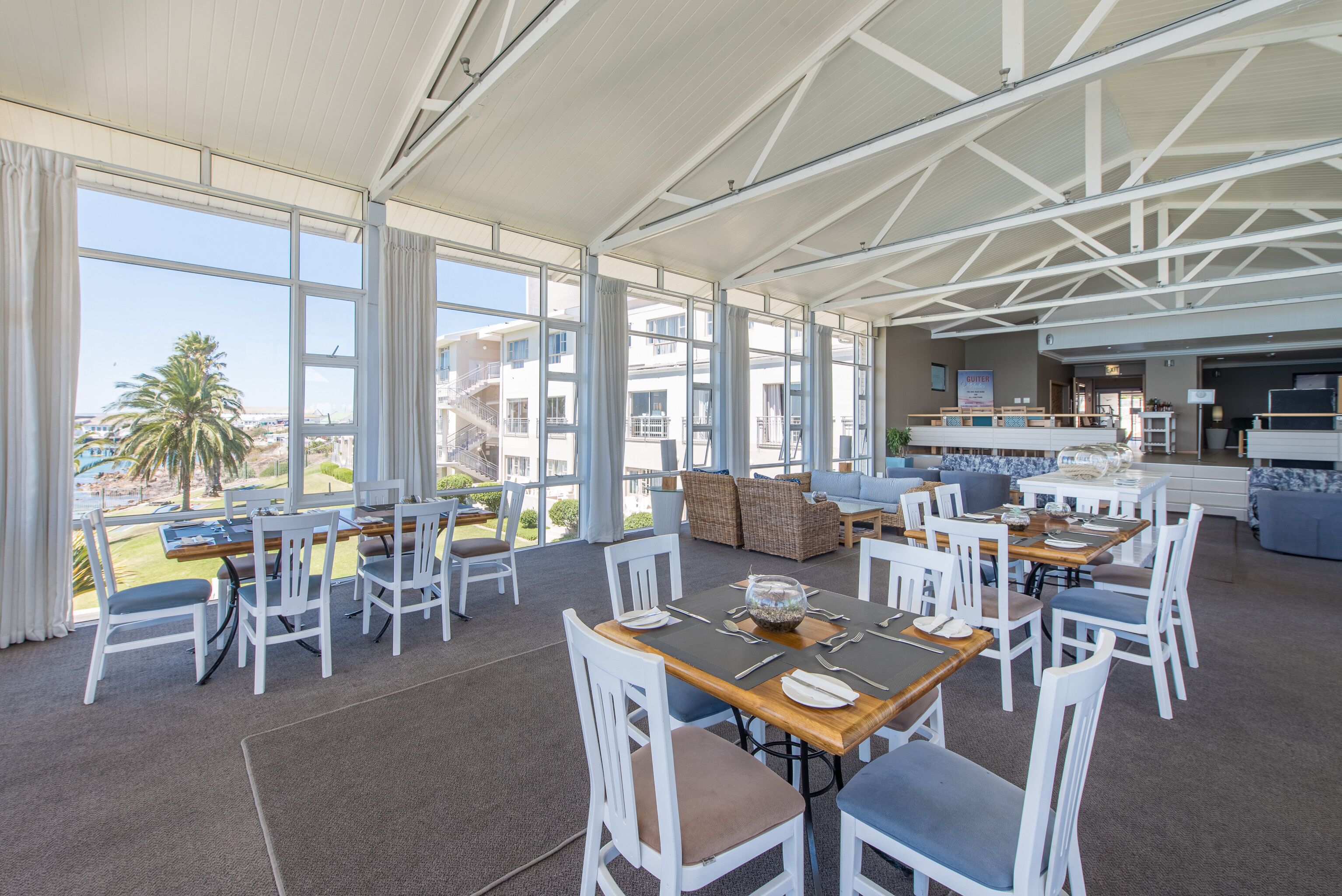 A spacious indoor dining area with multiple tables and chairs, large windows offering views of the outside, and a modern architectural ceiling design.