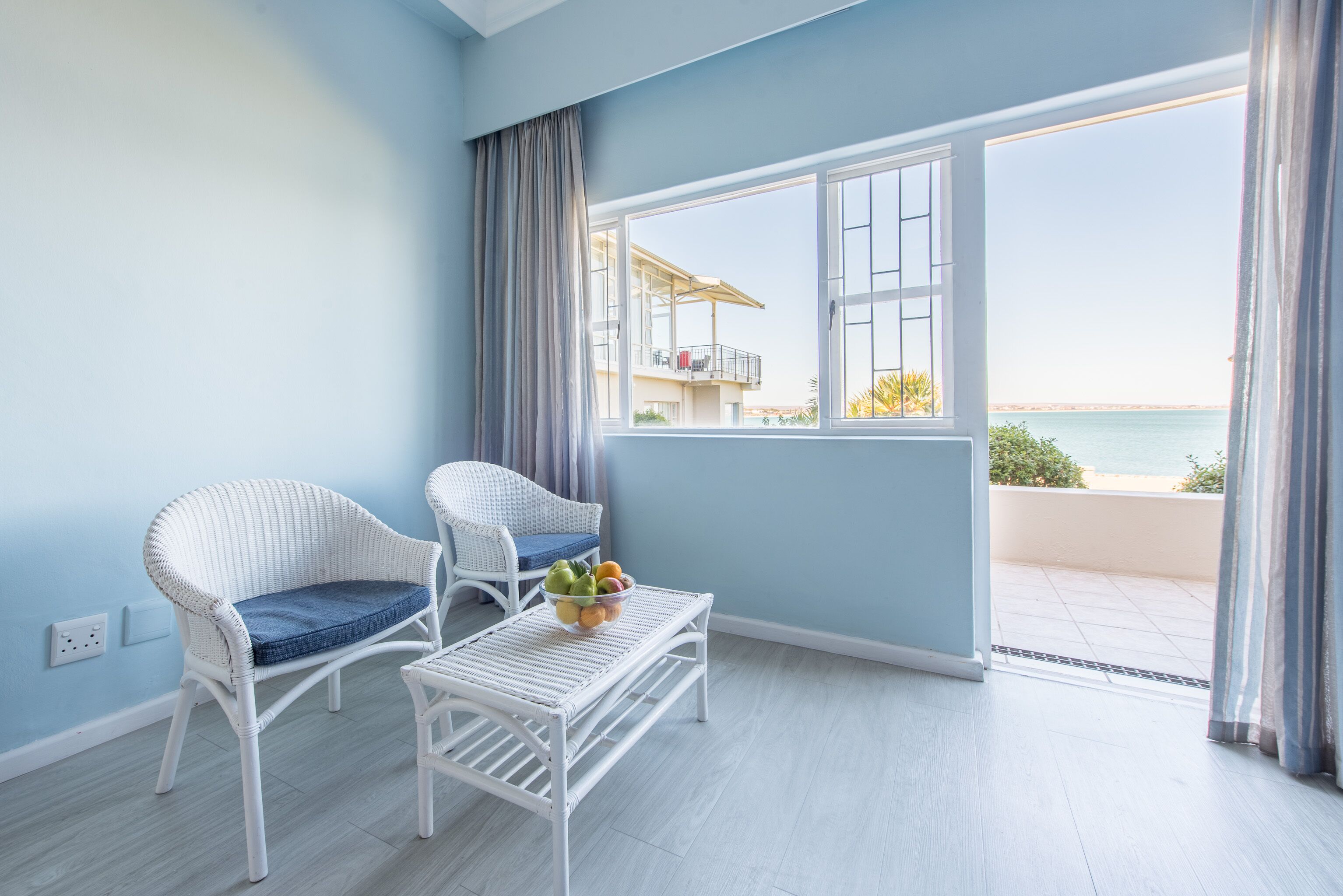 Une pièce avec deux fauteuils en osier, une petite table blanche sur laquelle se trouve un panier de fruits, et une grande fenêtre donnant sur la mer.