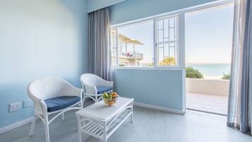 Une pièce avec deux fauteuils en osier, une petite table blanche sur laquelle se trouve un panier de fruits, et une grande fenêtre donnant sur la mer.